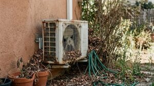Unidad exterior del aire acondicionado con hojas y objetos alrededor que dificultan la ventilación.