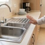 : Persona evitando tocar un fregadero metálico por posible riesgo eléctrico en la cocina.