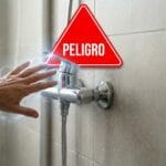 Mano recibiendo una descarga eléctrica al tocar un grifo de ducha con una señal de peligro en rojo en un baño alicatado